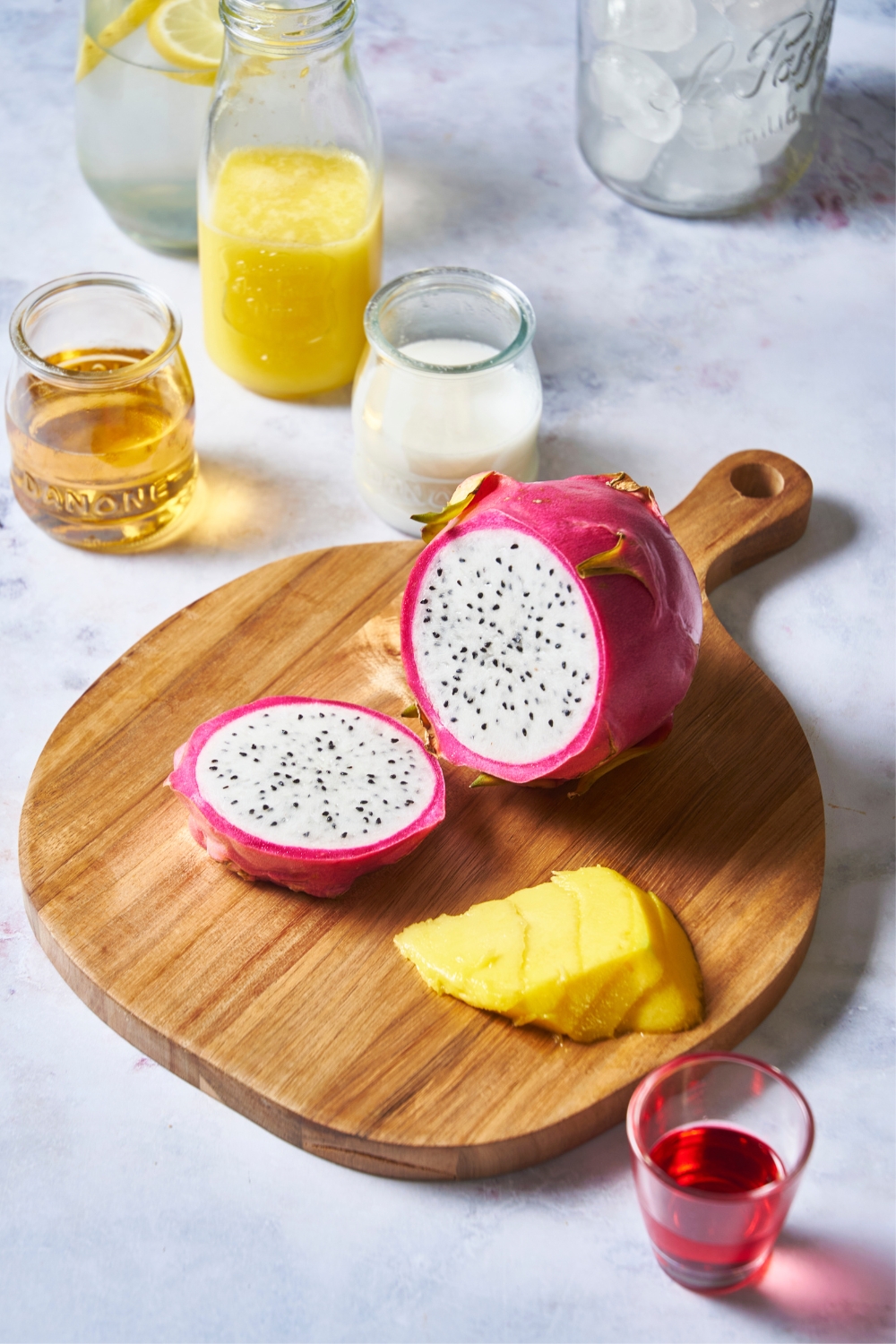A dragonfruit on a wooden cutting board. Small glasses with juice and a small shot glass with grenadine sit around to it.