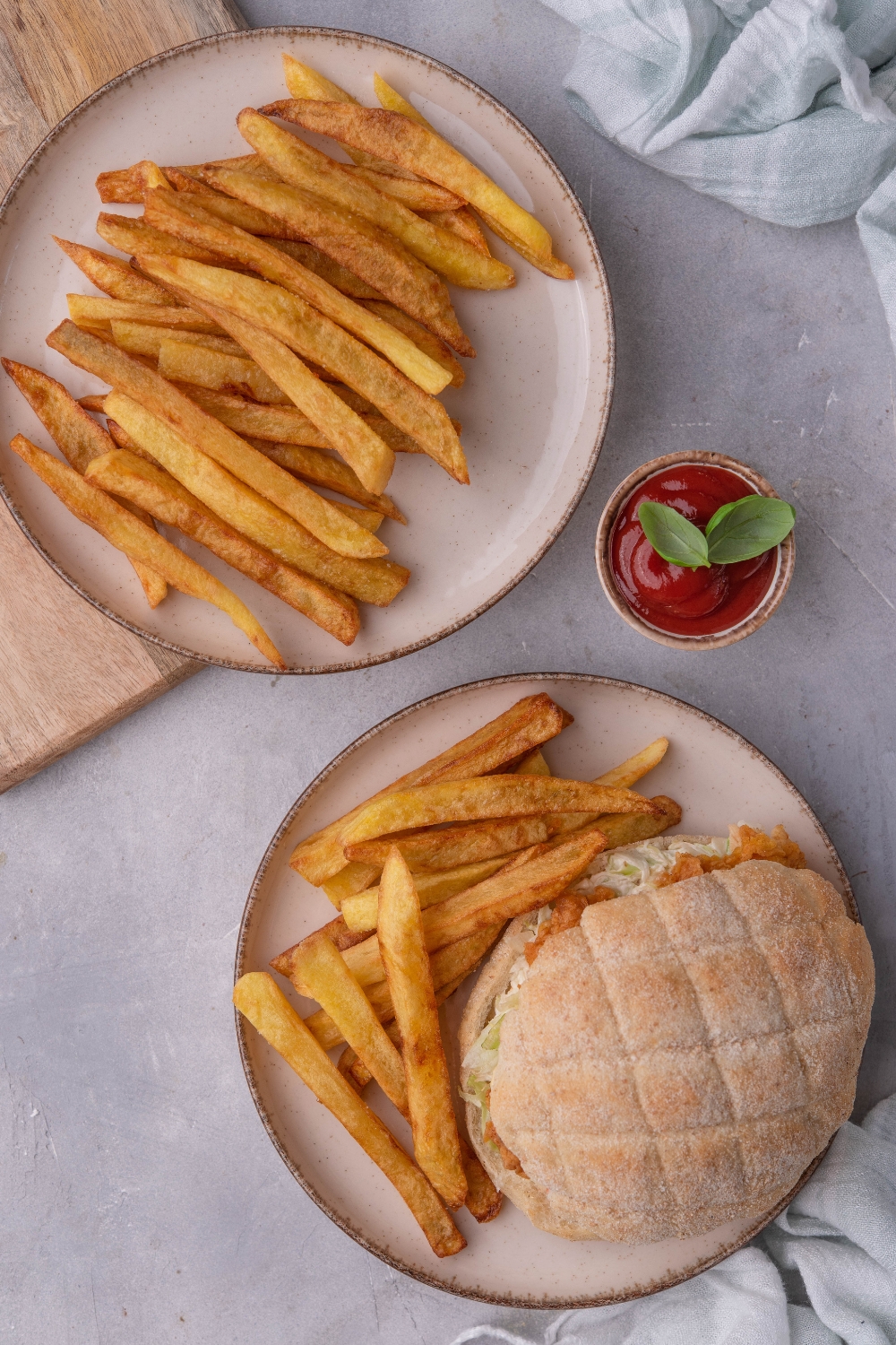 An overhead view of two plates, one with homemade french fries and another with fries and a sandwich on it. A small dipping cup with ketchup sits in the middle of it.