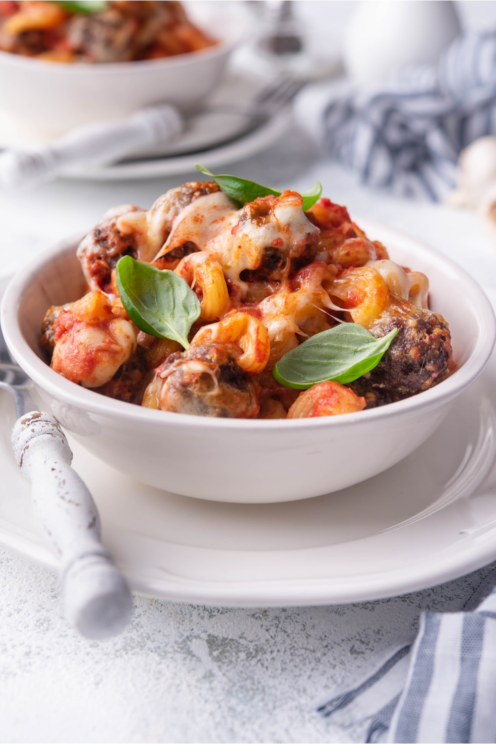 A white bowl of meatball casserole on a white plate with a fork on the plate. In the background there is another serving of casserole.