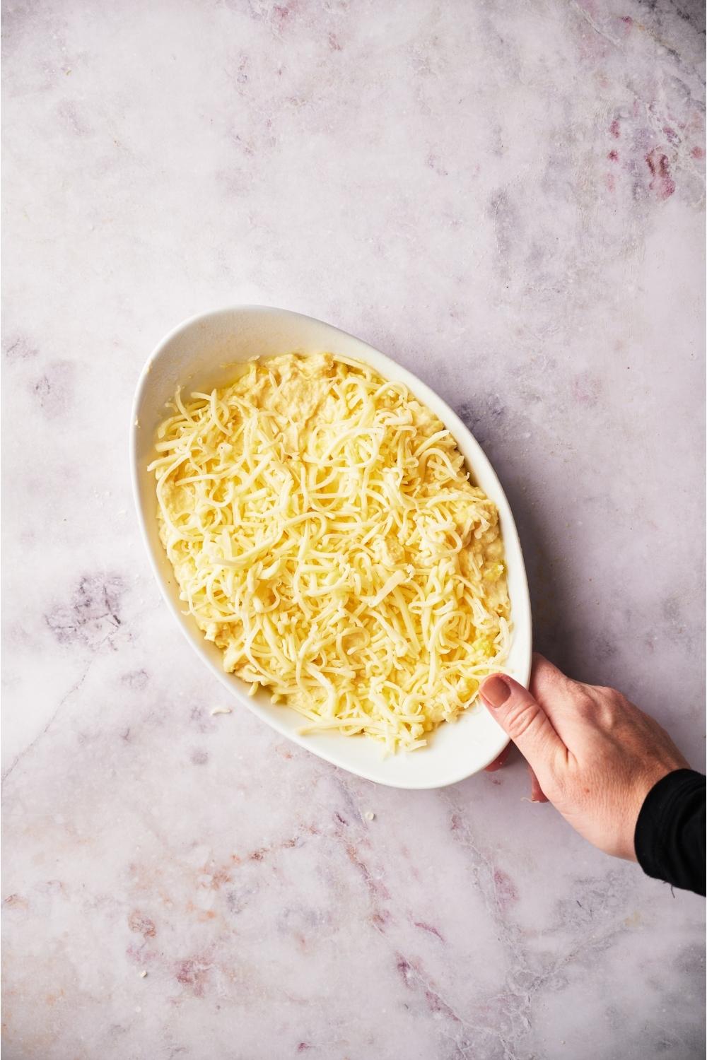 Hand grabbing white casserole dish with unbaked corn casserole topped with shredded cheese