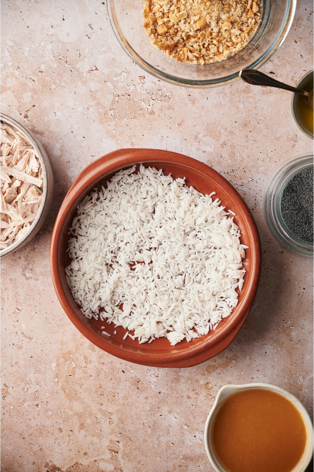 A red casserole dish with a layer of cooked white rice. Surrounding the dish there are bowls of ingredients including cracker crumbs, poppy seeds, shredded chicken, and broth.