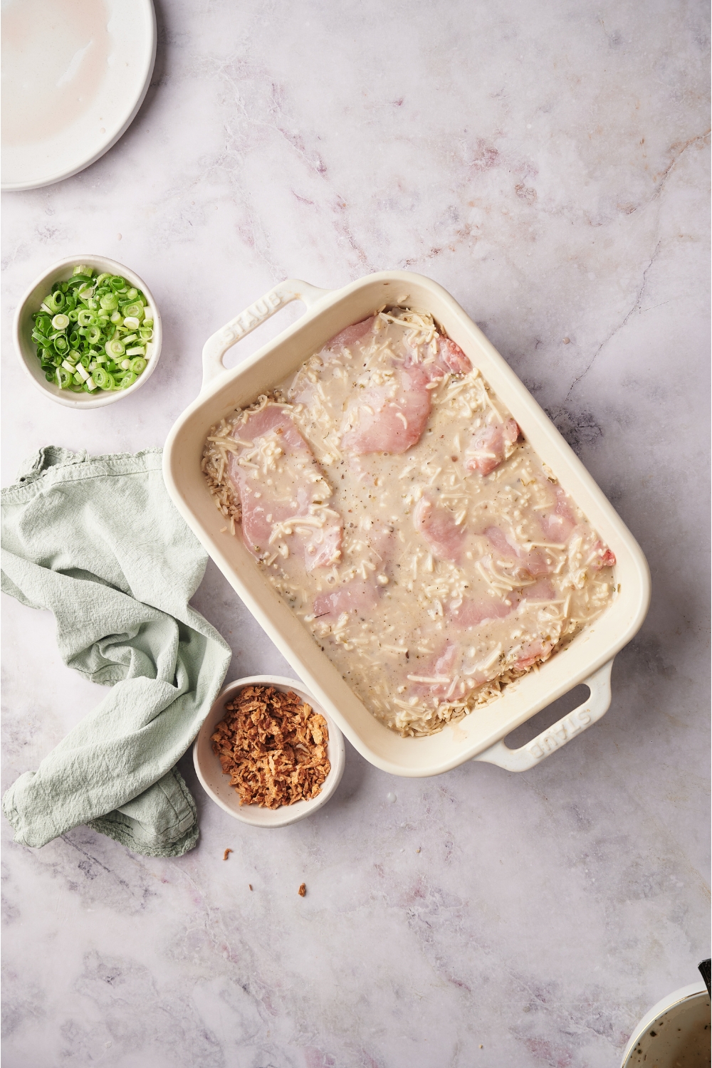 White baking dish filled with raw pork in a soup mixture with rice and cheese added. Next to the baking dish is a kitchen towel and bowls of green onions and fried onions.