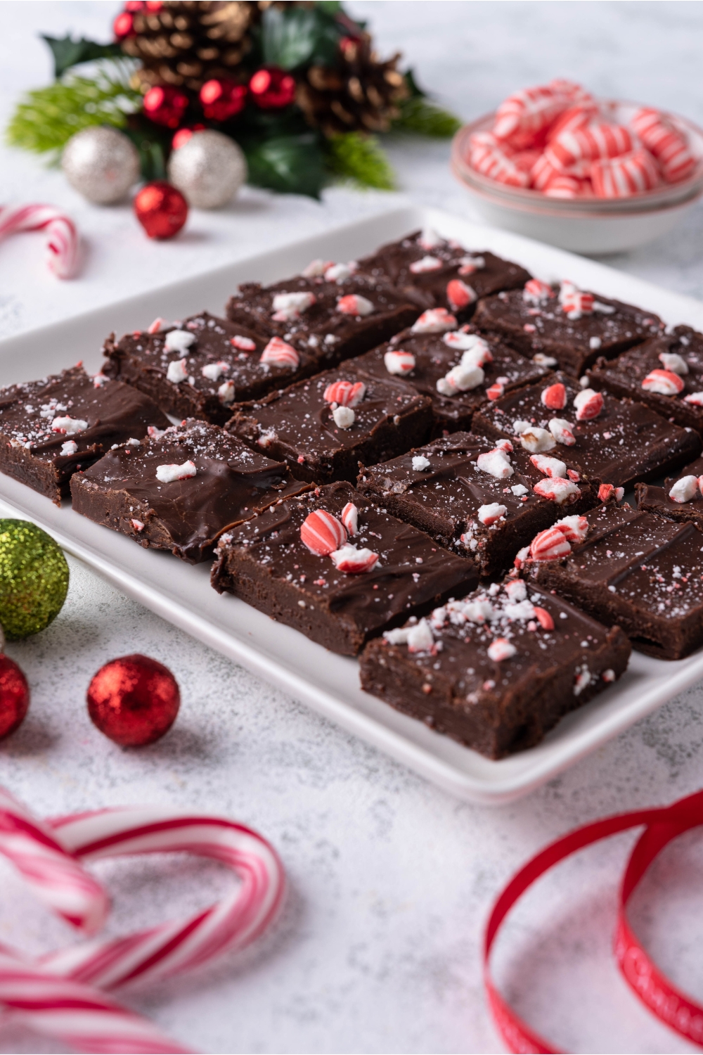 Peppermint fudge sliced into squares on a white serving tray with a bowl of peppermint candies and candy canes surrounding the serving tray.
