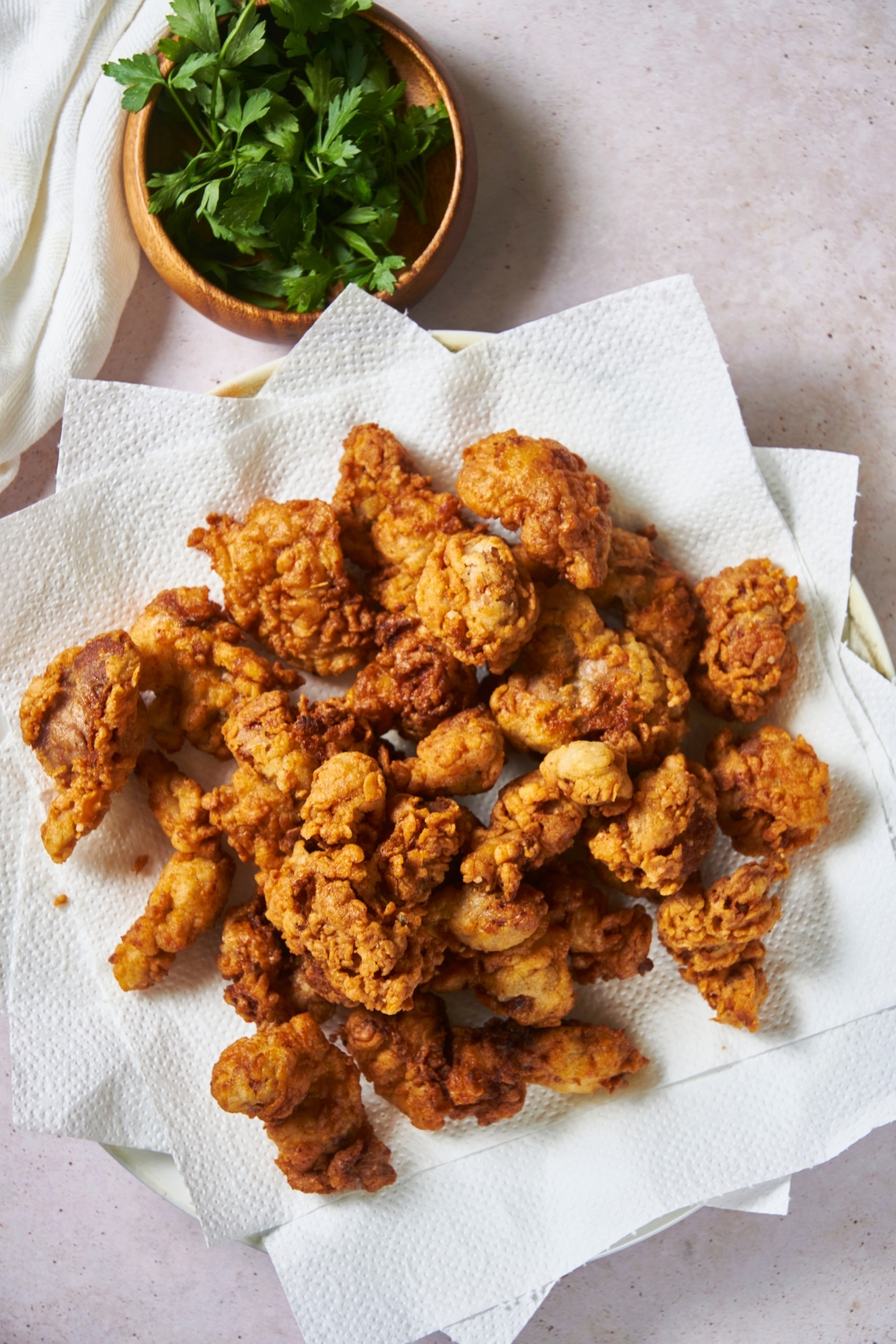A plate lined with paper towels and filled with fried chicken pieces. Next to the plate is a bowl of fresh herbs.