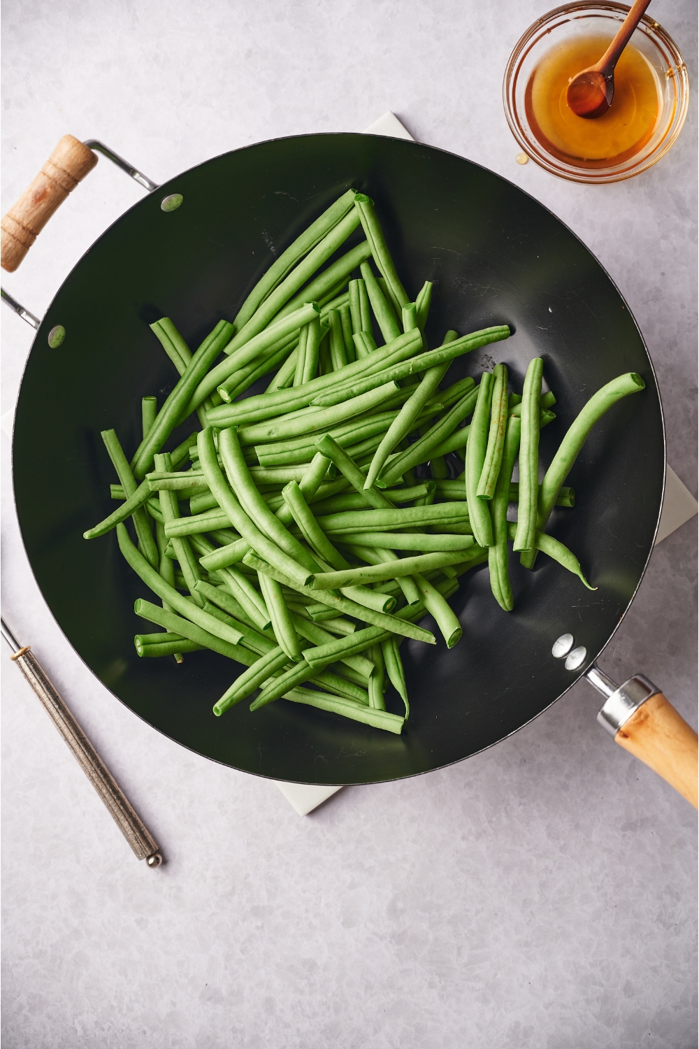 A black skillet filled with raw green beans. Next to the skillet is a small bowl of honey.