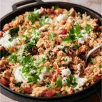 Black skillet on a wood board, filled with stuffed pepper casserole. There is a spoon in the skillet and the casserole is topped with sour cream and fresh herbs.