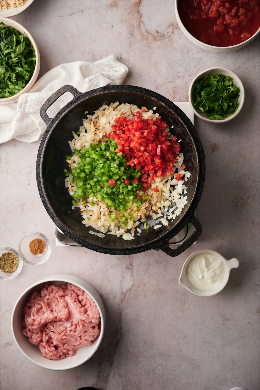Black skillet with diced onion, diced green peppers, and diced red peppers. The skillet is surrounded by ingredients including bowls of raw turkey, sour cream, parsley, and seasonings.