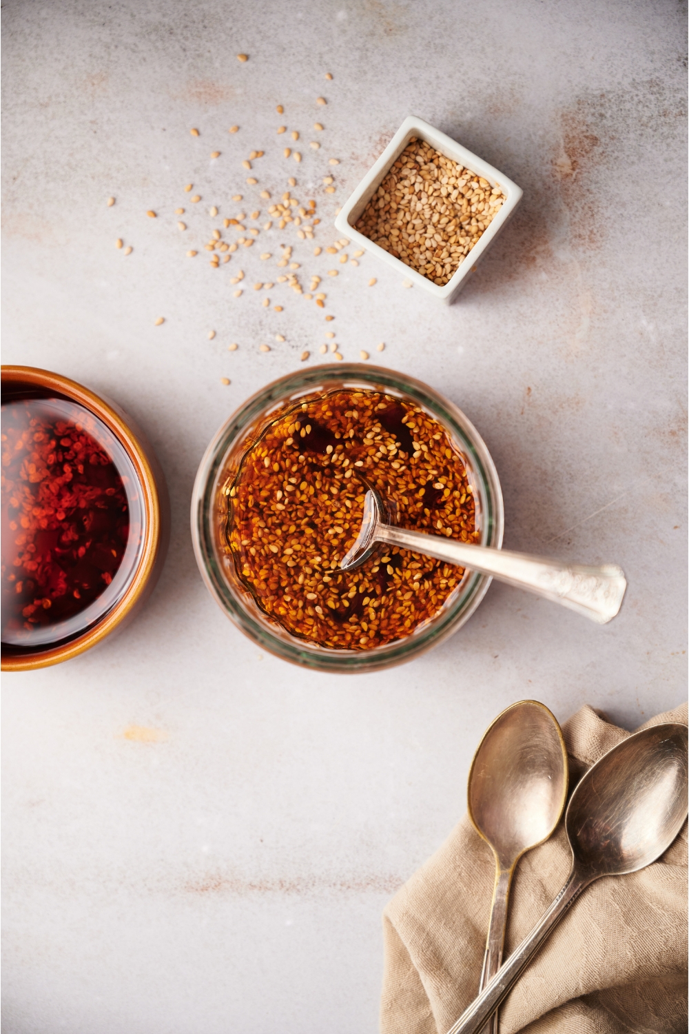 A clear bowl with sauce and a spoon in the bowl. Next to the bowl there are 2 spoons and bowls of sesame seeds and chili oil.