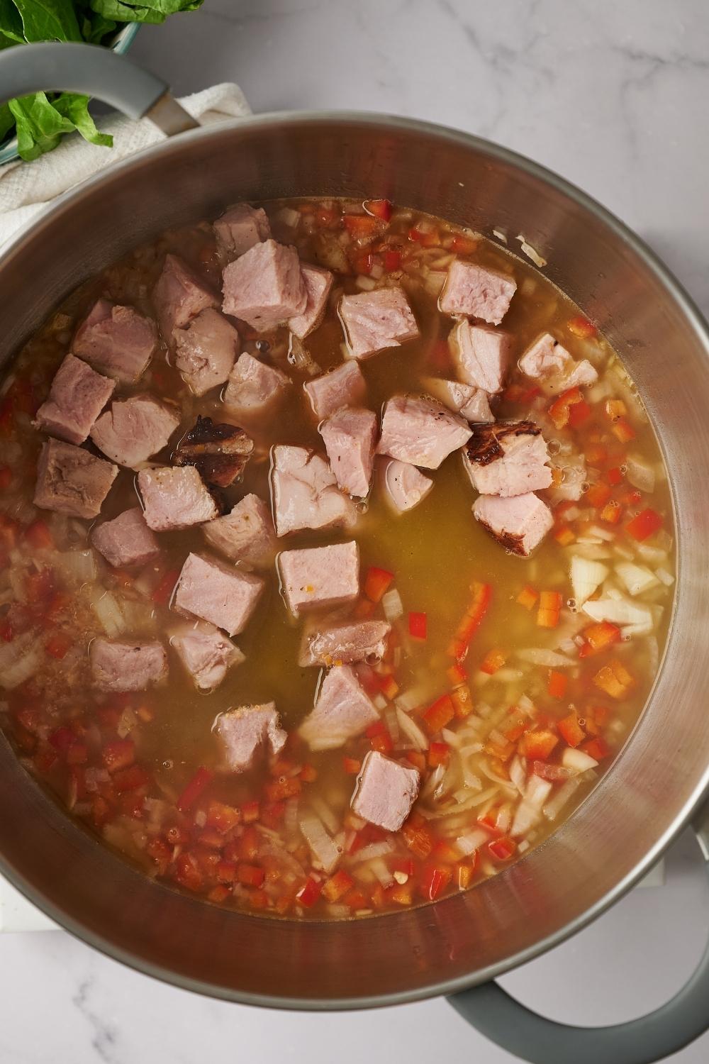 A large hot pot with diced onion, peppers, and cubed turkey ham in it. Beef broth has just been added to it.
