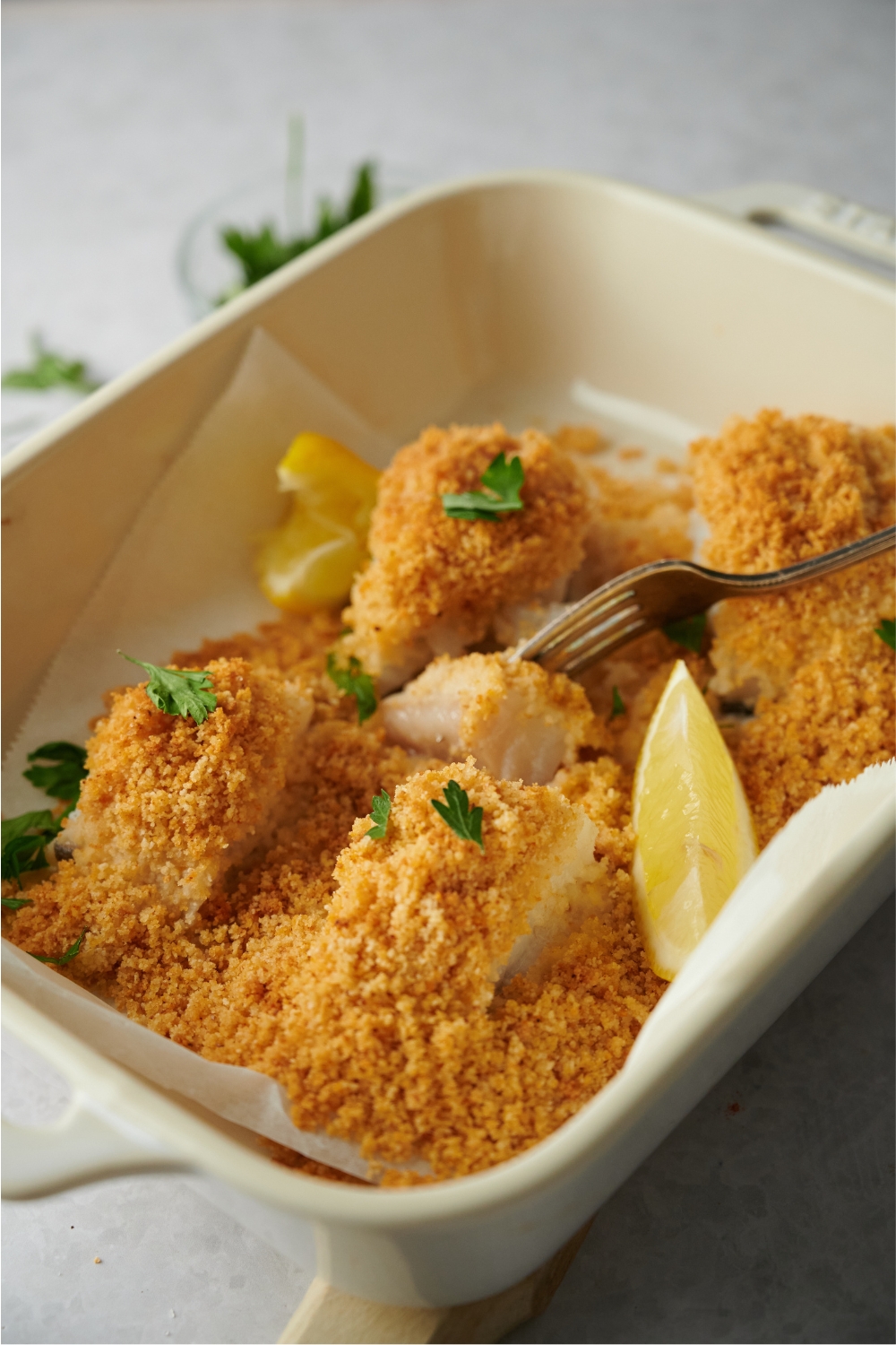A baking dish filled with breaded haddock fillets. A fork has a piece of the one fillet in the prongs.