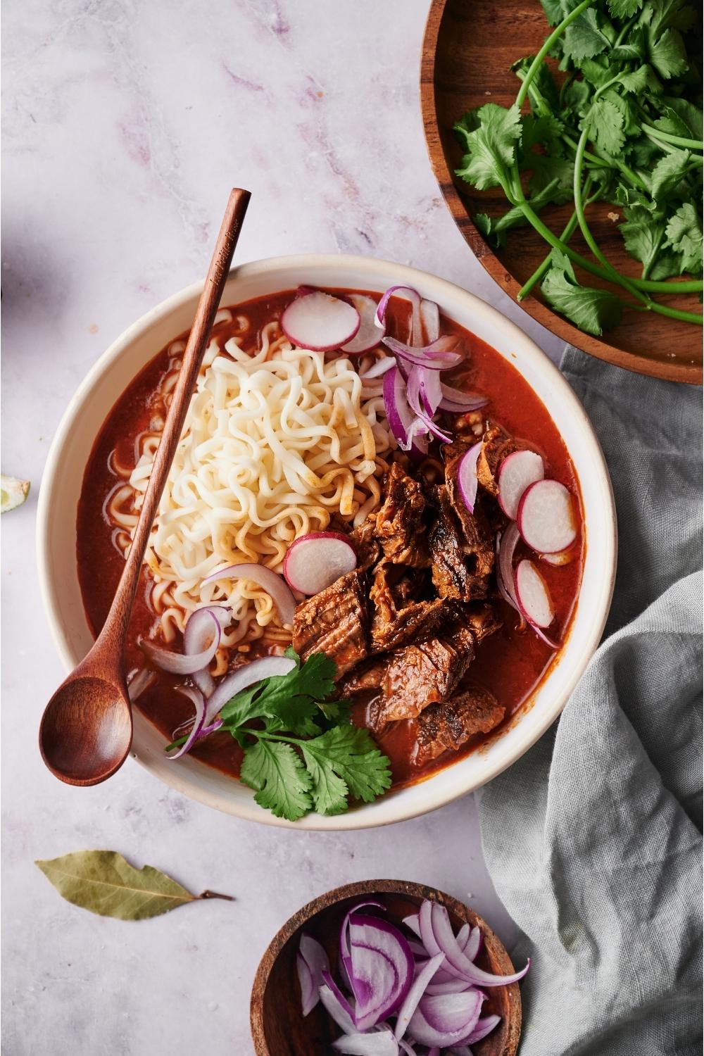 Birria ramen in a white bowl with a spoon, surrounded by an assortment of toppings