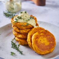 Sweet potato patties stacked on top of each other with some off to the side, topped with an herb mayo and additional fresh herbs.