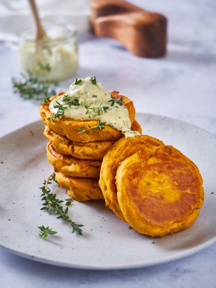 Sweet potato patties stacked on top of each other with some off to the side, topped with an herb mayo and additional fresh herbs.