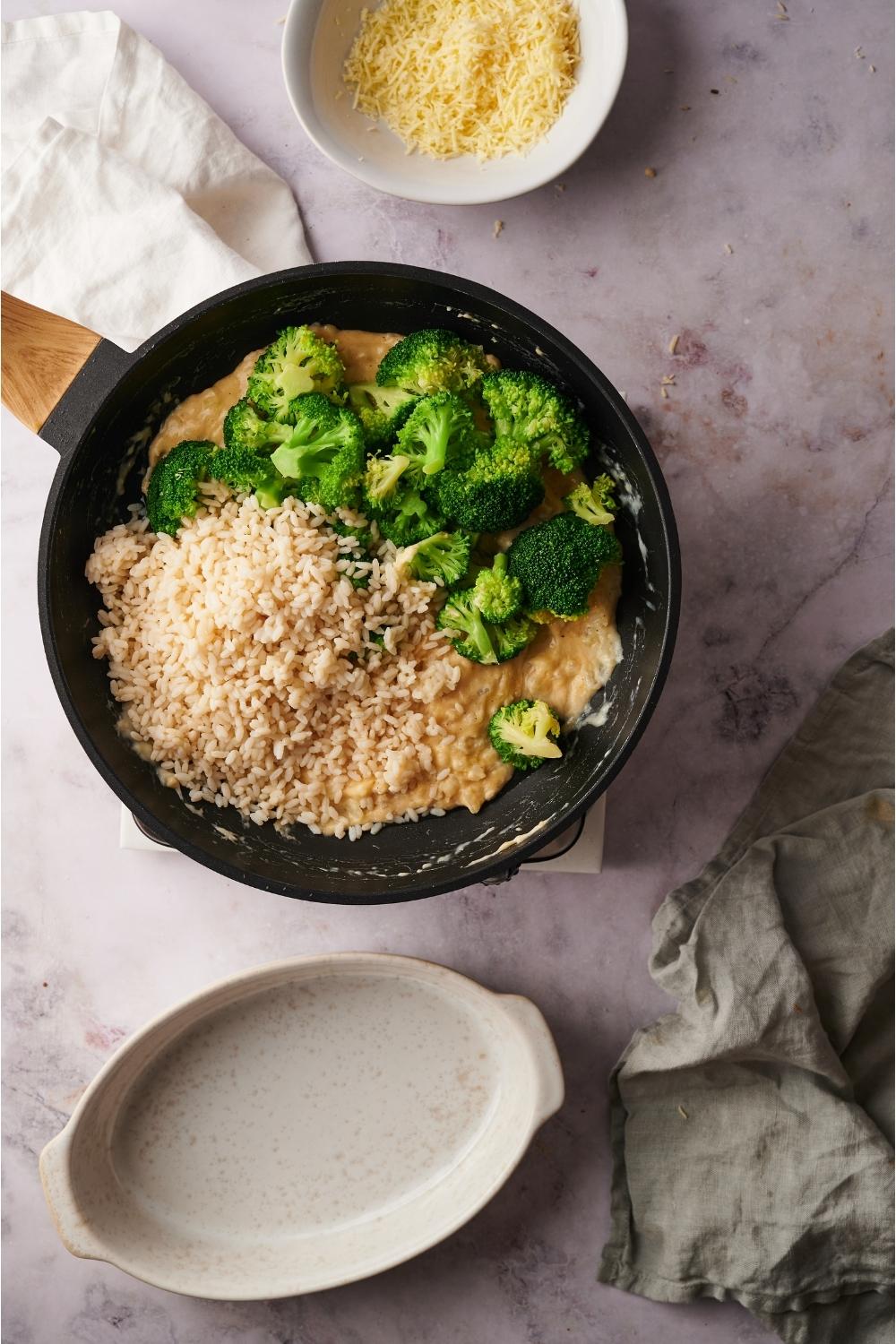 Skillet with rice and broccoli freshly added to a cheesy sauce to make broccoli rice casserole, next to an empty plate and a bowl of shredded cheese.