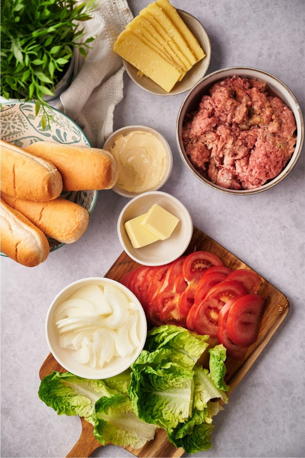 Lettuce, onion, and tomato on a wooden cutting board. Behind it is part of a bowl with for sub rolls in a bowl with ground beef in it, and a bowl of slices of cheese in it.