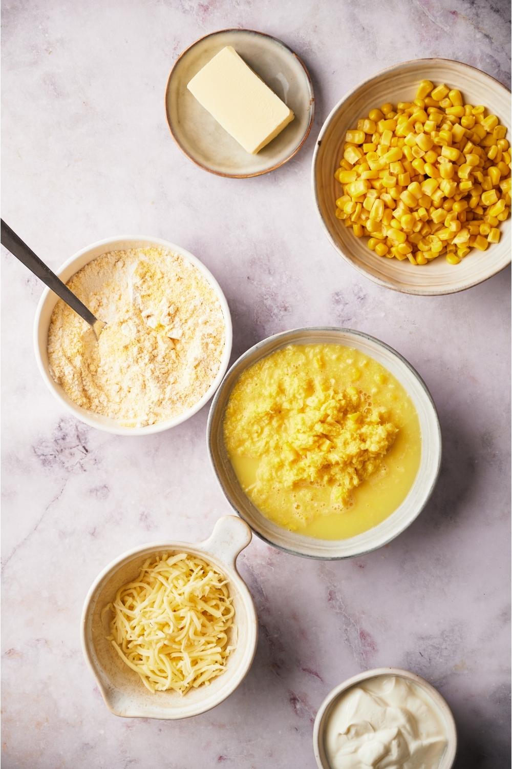 An assortment of ingredients for Paula Deen's corn casserole including bowls of creamed corn, fresh corn, corn muffin mix, shredded cheese and butter