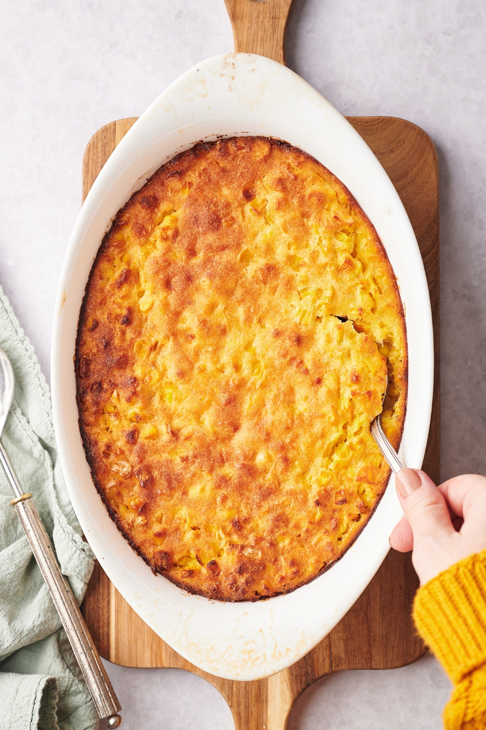 A casserole dish with cooked corn souffle. A hand is scooping a bit of it with a spoon.