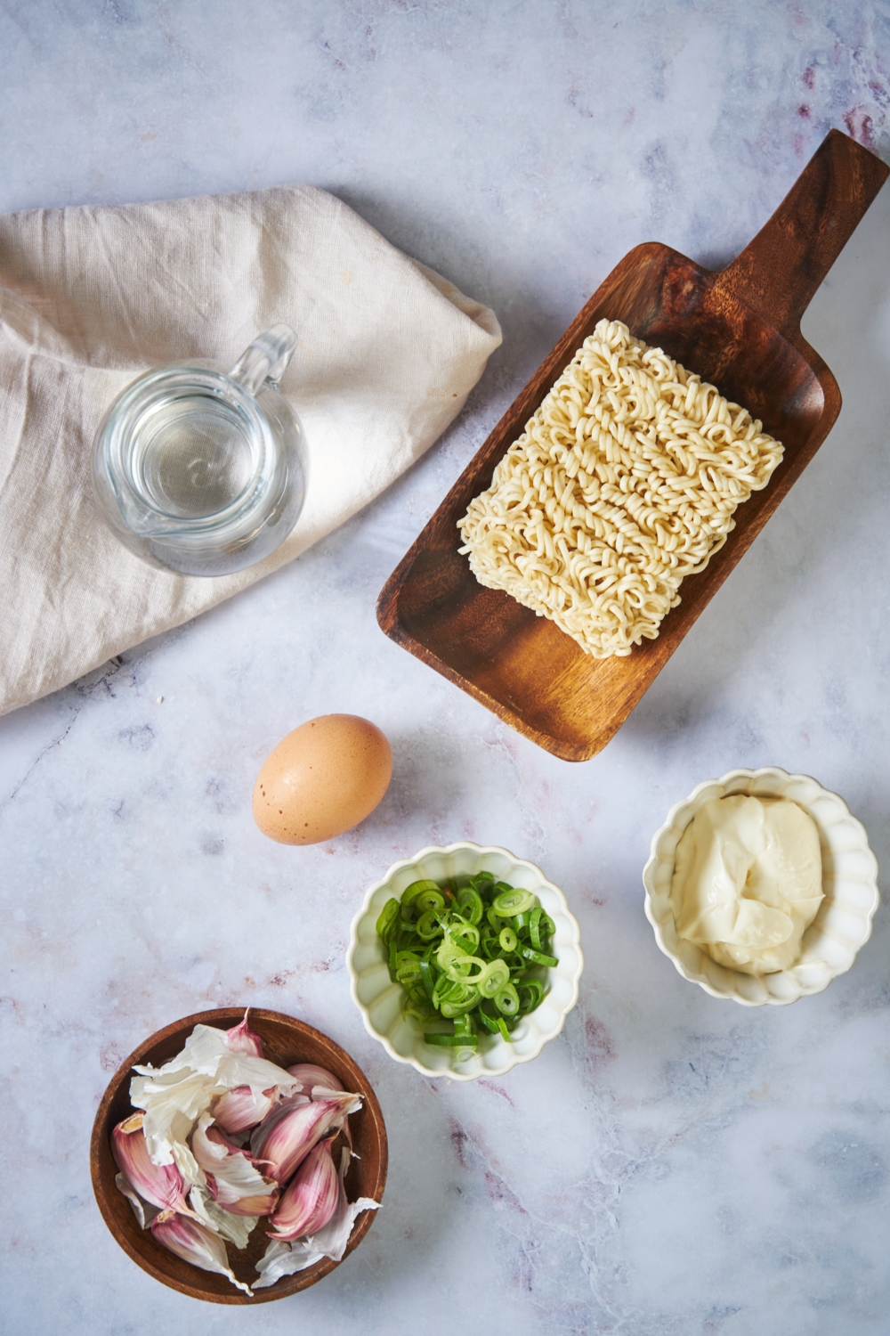 A block of ramen on a wood board, a bowl of garlic, an egg, bowl of scallions, and a bowl of mayo all on a grey counter.