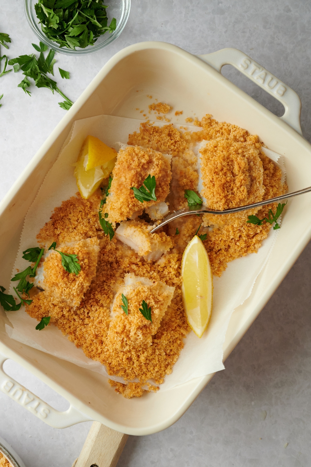 A fork with a piece of haddock on it in a baking dish that is lined with parchment paper with four breaded haddocks in it.