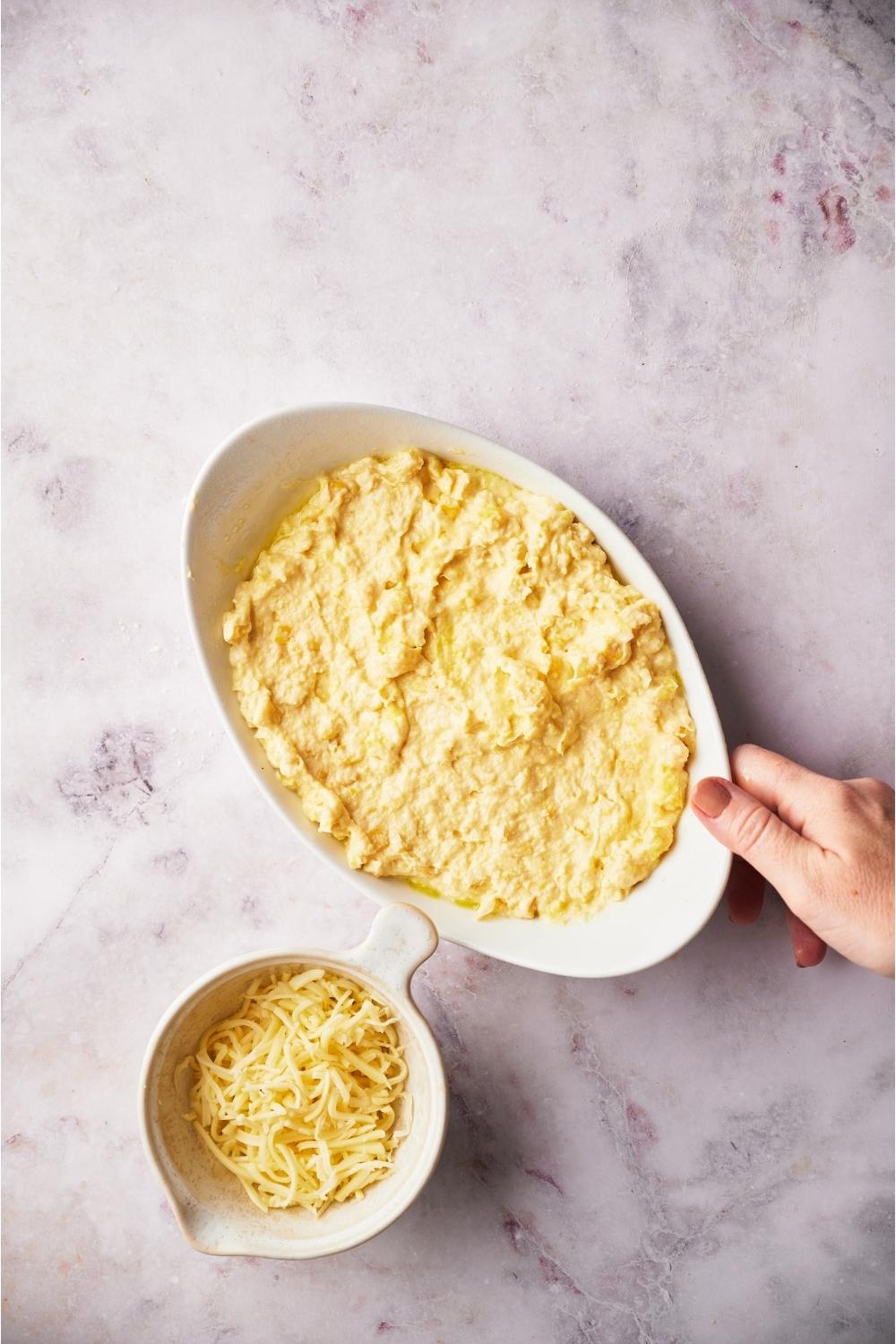 Hand grabbing white casserole dish with unbaked corn casserole next to a small bowl of shredded cheese