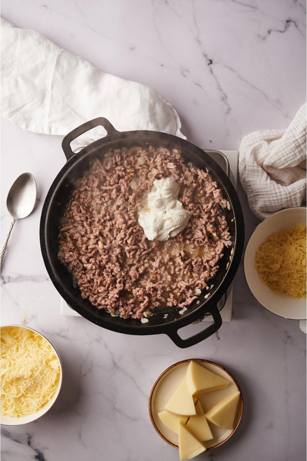 A skillet with cooked ground beef and cream cheese freshly added, the skillet is next to plates of shredded and sliced cheese.