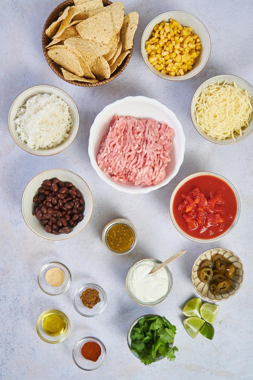 An assortment of ingredients for ground turkey casserole including bowls of raw ground turkey, tomato sauce, cheese, corn, salsa, beans, rice, and tortilla chips