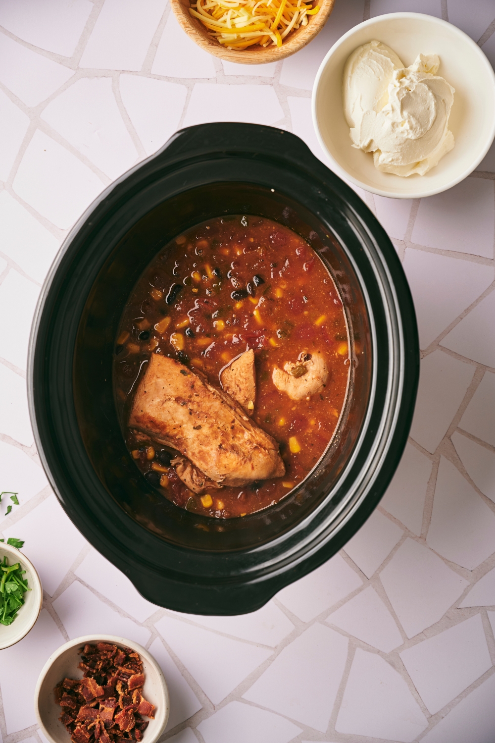 A cooked chicken breast, corn, and black beans in a broth in a slow cooker. Behind it is a bowl of cream cheese.