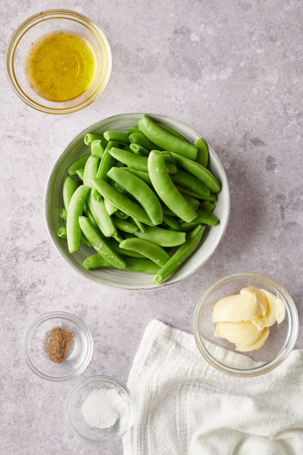 A countertop with a bowl of fresh sugar snap peas, oil, butter, salt, and pepper.