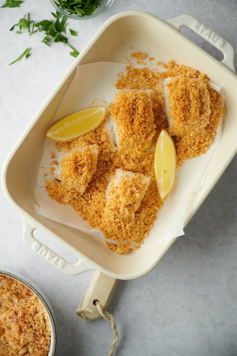 Two lemon wedges and four haddock fillets topped with bread crumbs on top of parchment paper in a baking dish.