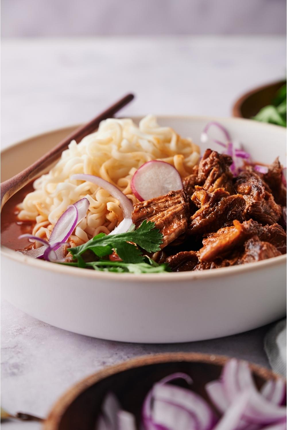 Birria ramen in a white bowl with a spoon, surrounded by an assortment of toppings