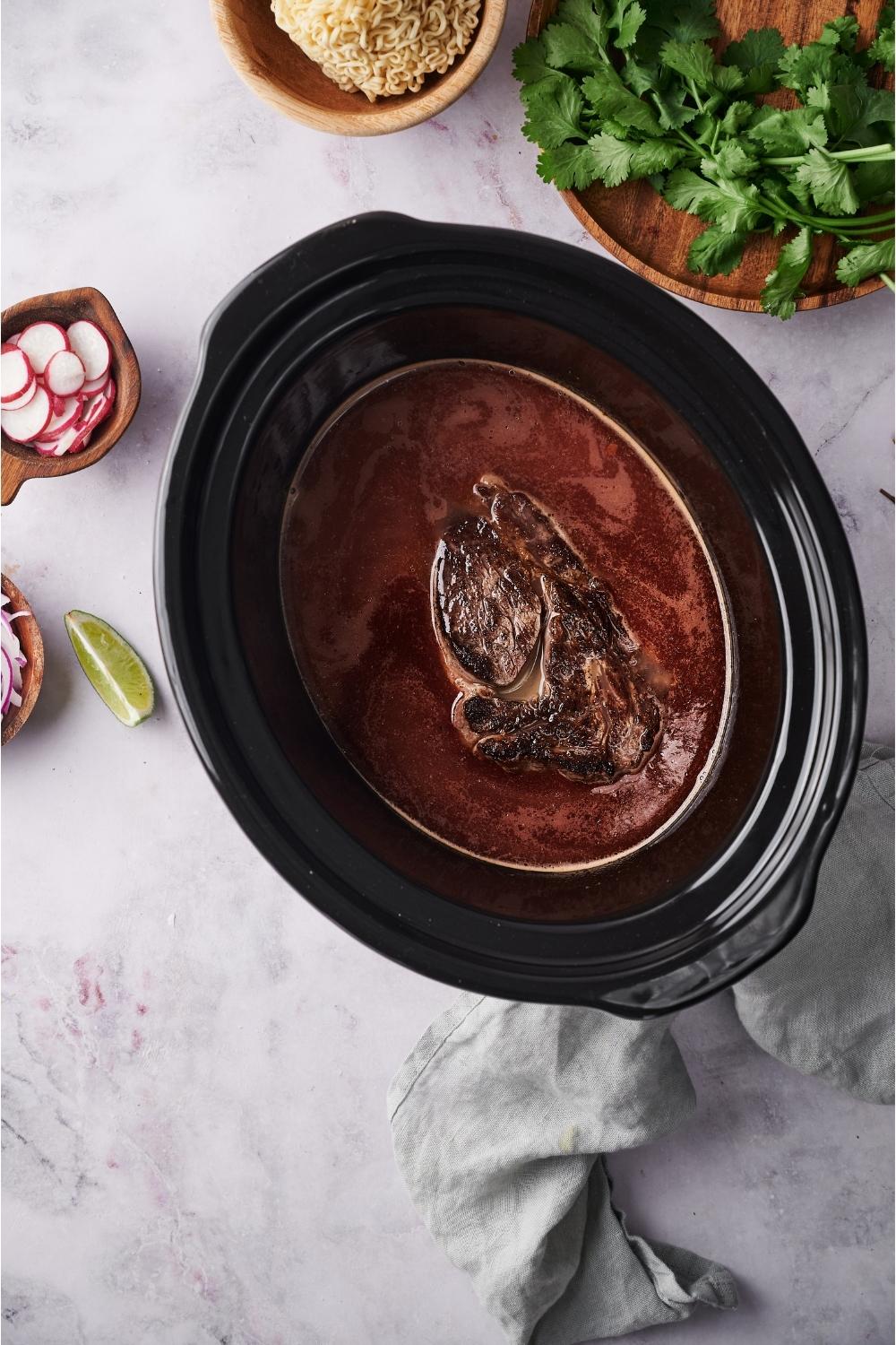 Crockpot with seared beef and beef broth, surrounded by ingredients for birria ramen