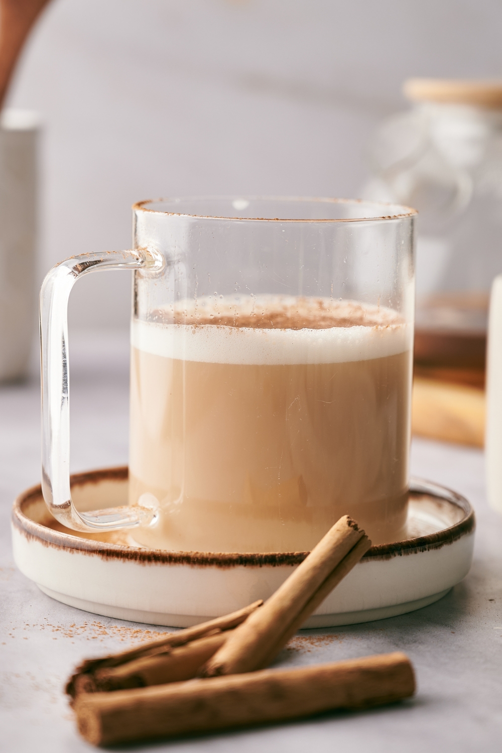 A glass that is filled with a chai tea latte. There are cinnamon sticks in front of the latte.