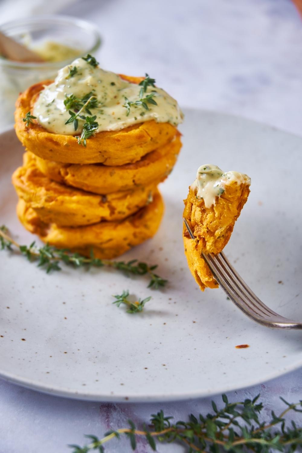 Sweet potato patties stacked on top of each other, topped with herb mayo. A fork is holding a piece of a sweet potato patty over top the plate.