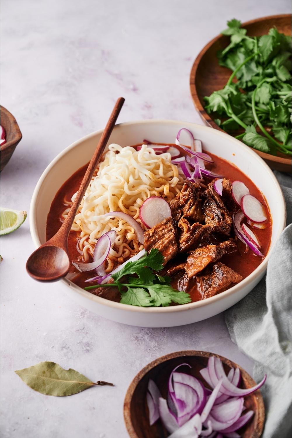 Birria ramen in a white bowl with a spoon, surrounded by an assortment of toppings