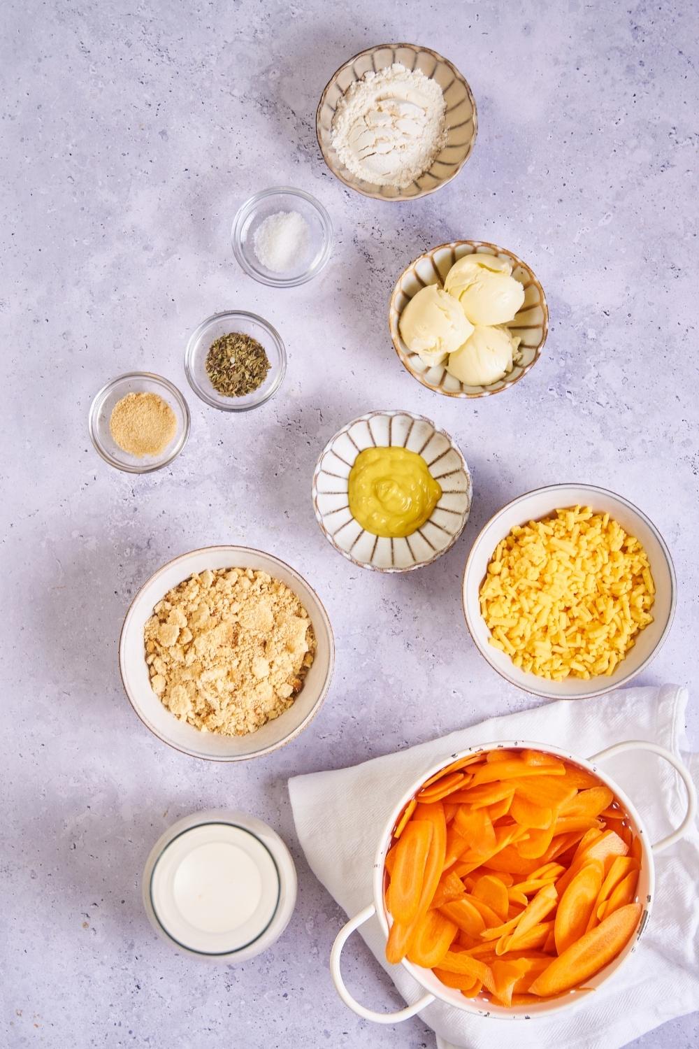 An assortment of ingredients for carrot casserole including bowls of carrots, mustard corn, cheese, flour, and spices.