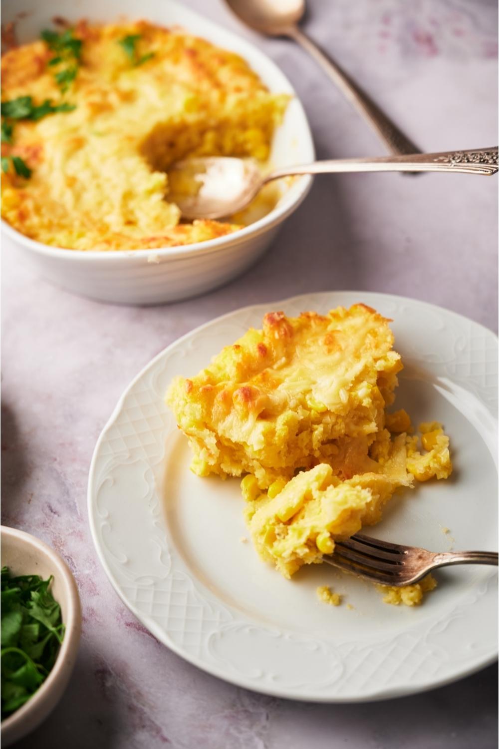 A serving of Paula Deen's corn casserole with a fork on the plate and the casserole dish in the background