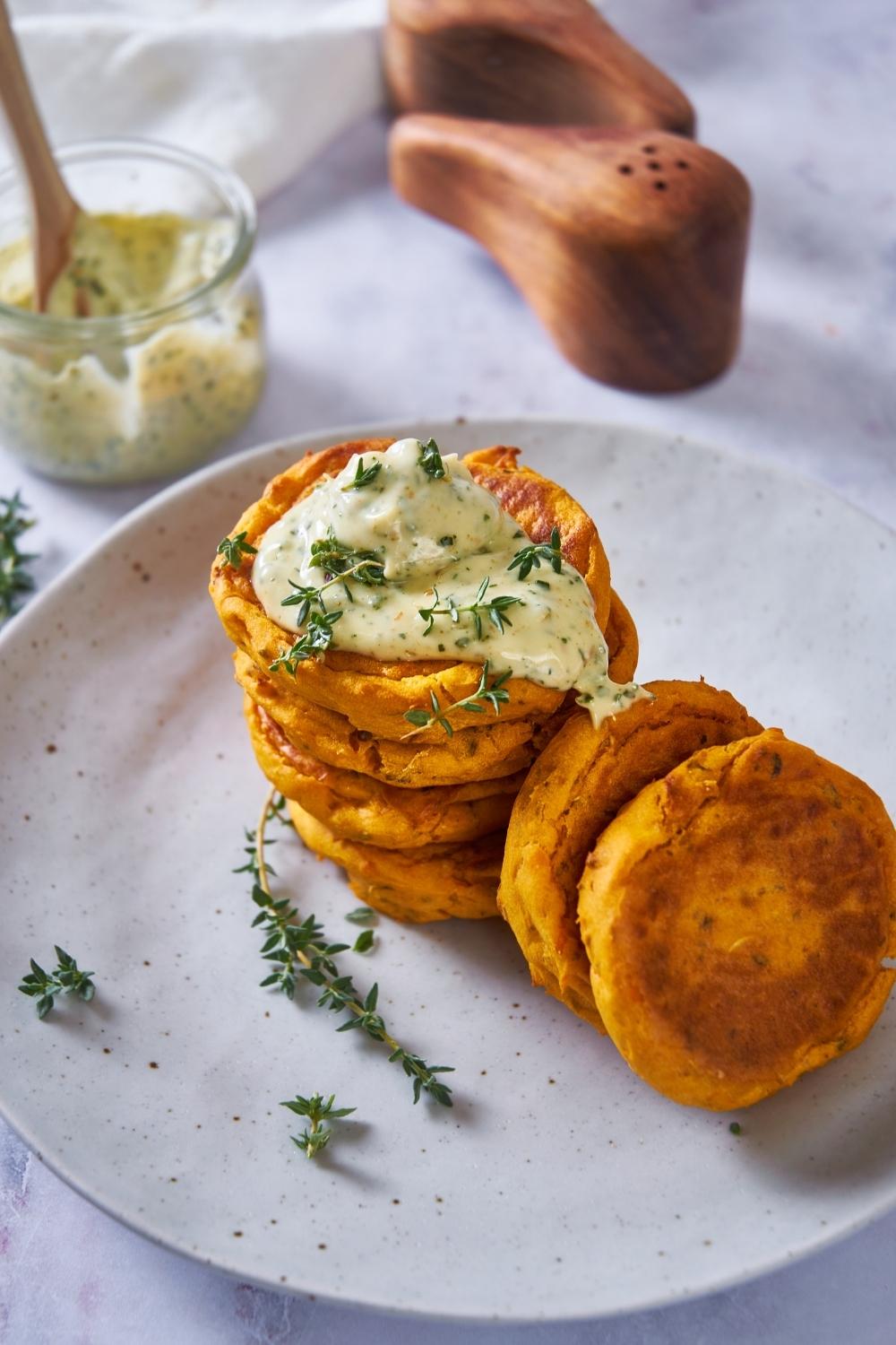 Sweet potato patties stacked on top of each other with some off to the side, topped with an herb mayo and additional fresh herbs. A jar of herb mayo is in the background next to a salt and pepper shaker.