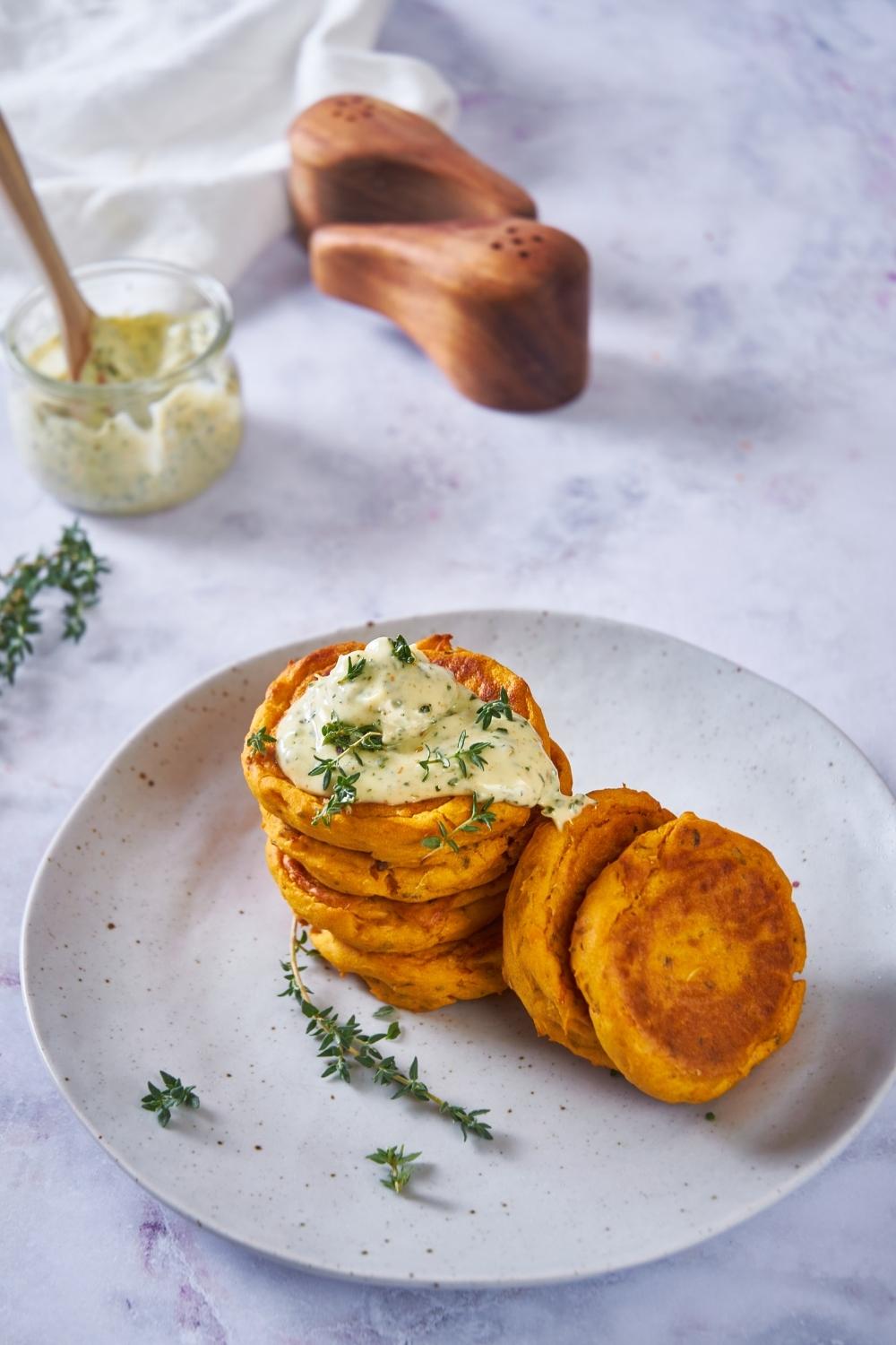 Sweet potato patties stacked on top of each other with some off to the side, topped with an herb mayo and additional fresh herbs. An empty jar of herb mayo is in the background next to a salt and pepper shaker.
