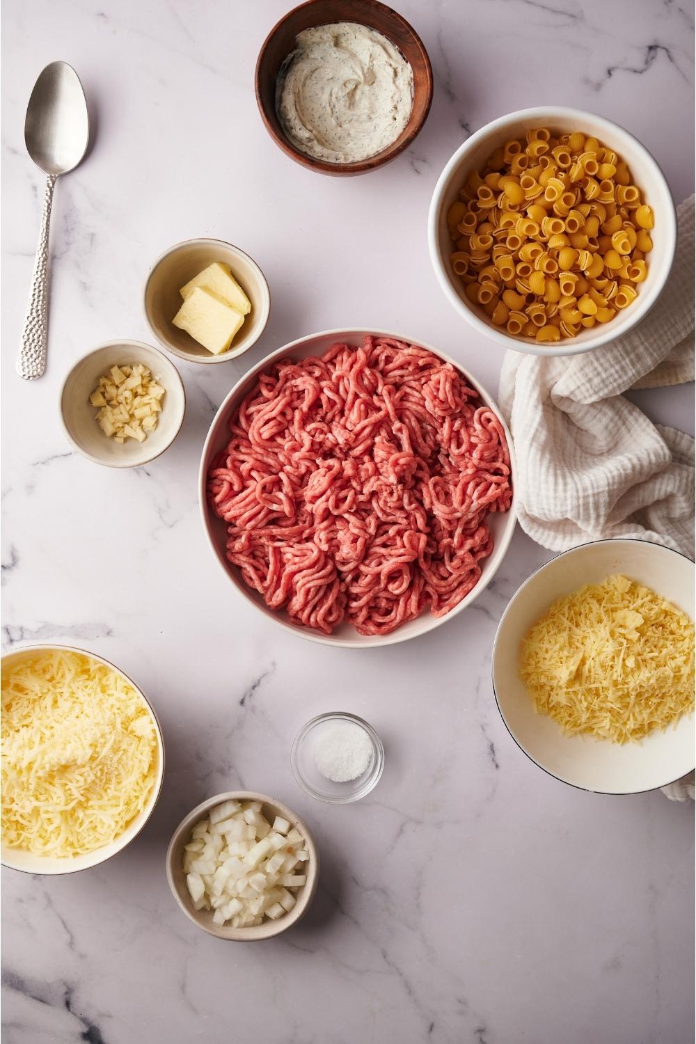 An assortment of ingredients for Philly cheesesteak casserole including bowls of raw beef, cheese, and uncooked pasta noodles.
