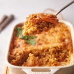 A casserole dish with shrimp casserole, a spoon is holding a large scoop over the dish.