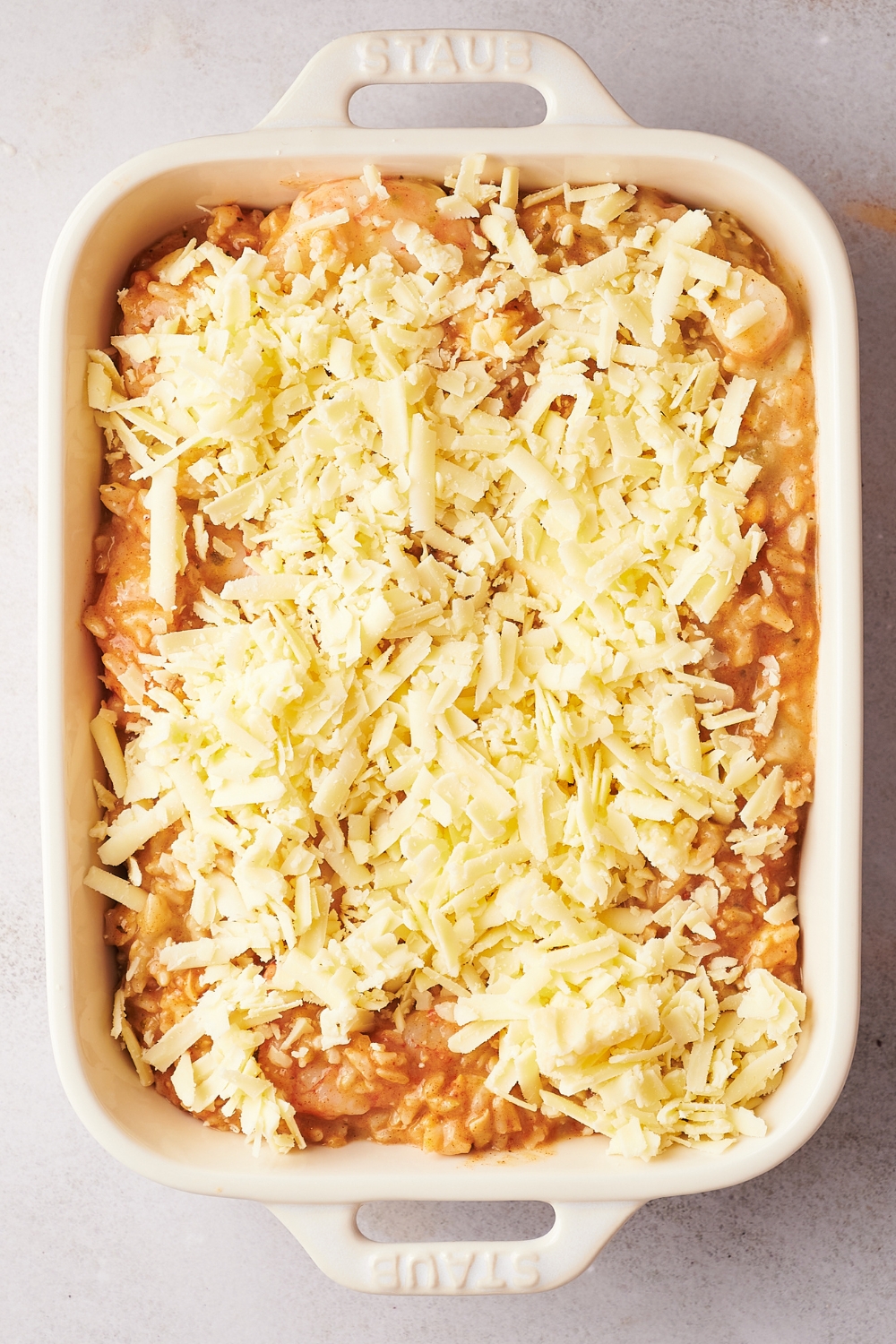 A casserole dish with uncooked shrimp casserole sprinkled with shredded cheese.
