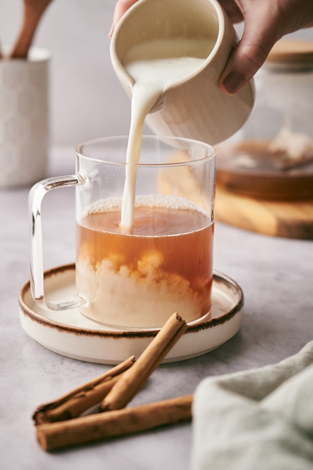 A clear mug with chai tea in it. A hand is pouring a small pitcher with milk into the mug.