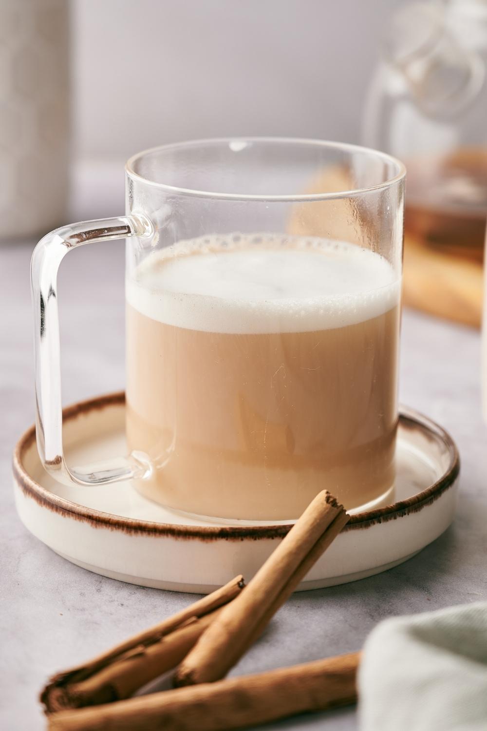 A clear mug with chai tea latte in it sitting on a ceramic saucer. Cinnamon sticks garnish the dish.