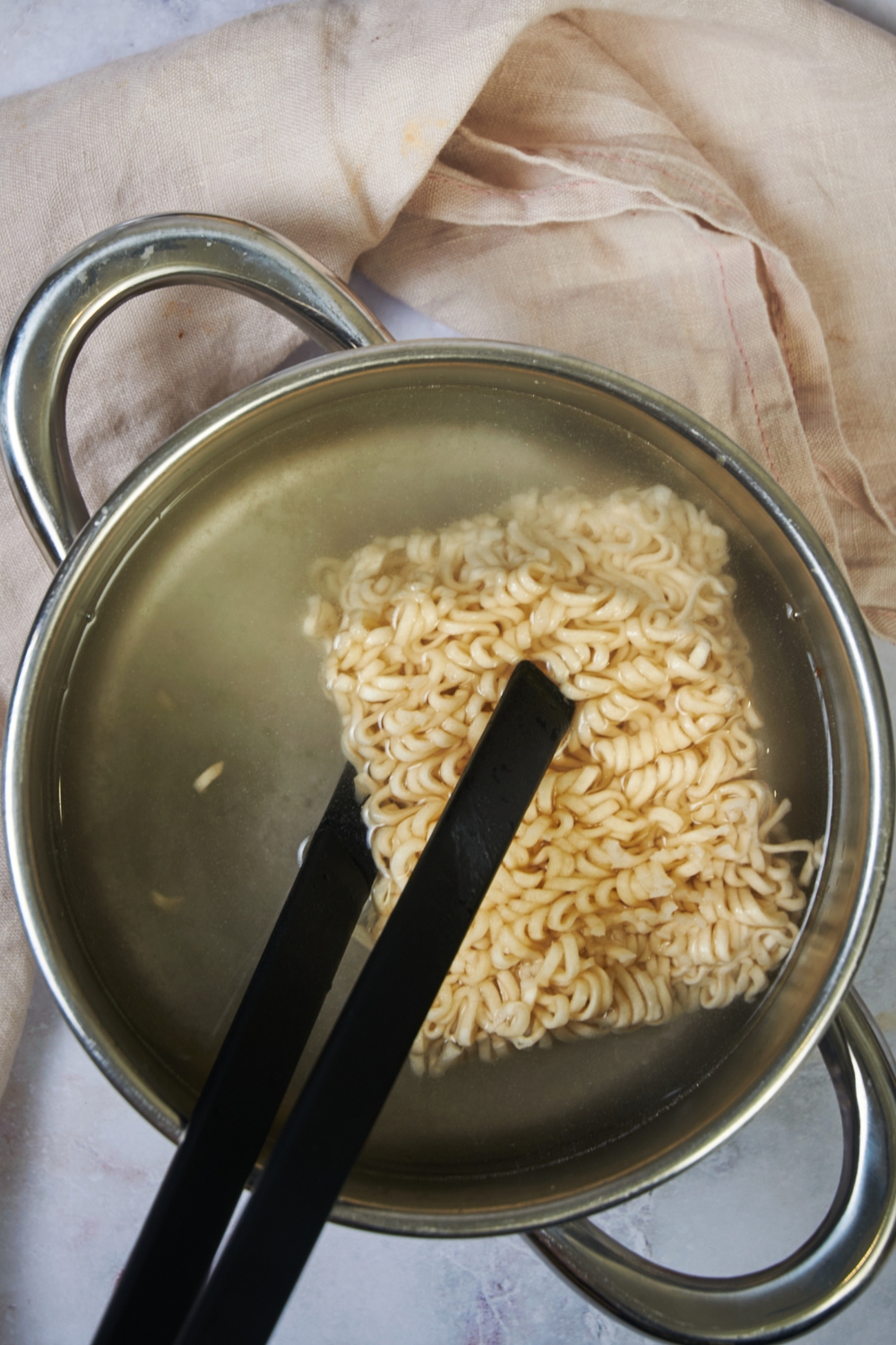 Tongs holding a block of ramen in a pot of water.