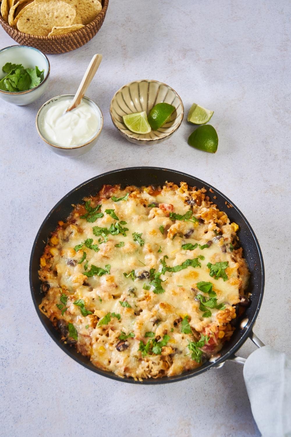 A black skillet filled with ground turkey, melted cheese, and cilantro. The skillet is next to bowls of lime wedges, sour cream, cilantro, and tortilla chips