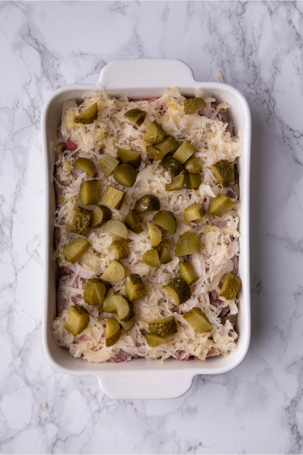 Layers of pickles, sauerkraut, cheese and pastrami in a white baking dish.