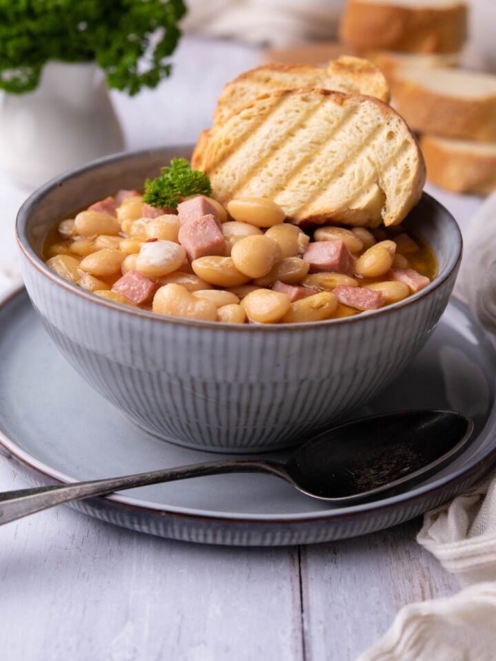 Butter beans with diced ham and two slices of toast on a blue bowl on top of a blue plate, with a spoon on the plate.