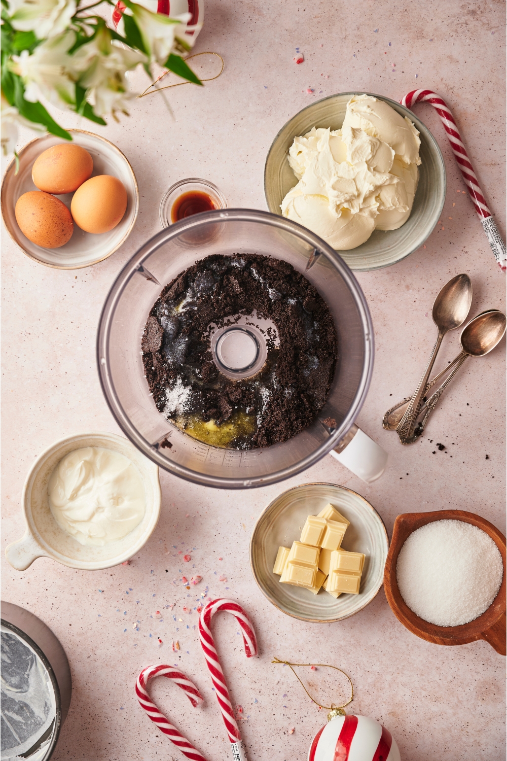 Food processor with dark chocolate crumbs and melted butter, surrounded by ingredients including bowls of white chocolate, cream cheese, eggs, sugar, and vanilla extract.