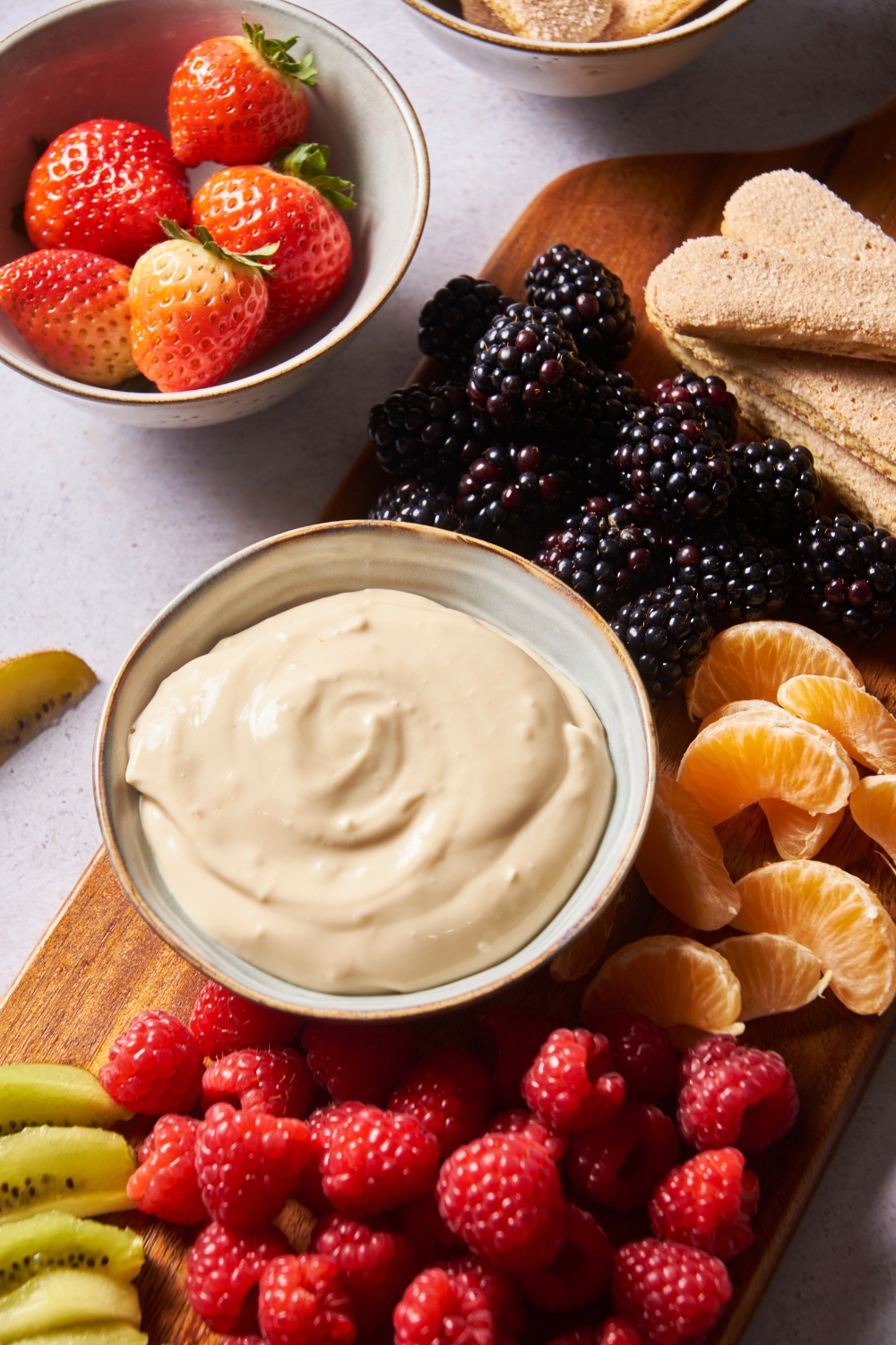 Cream cheese dip in a serving bowl on a wood board filled with fresh fruit.