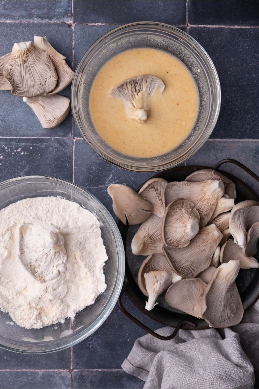 A plate of oyster mushrooms next to two bowls, one with a dry flour mixture and one with a wet mixture. One mushroom is in the wet mixture and one mushroom is in the dry mixture.